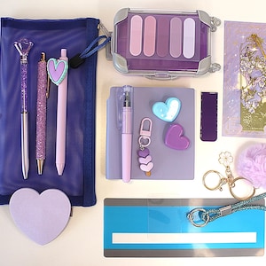 May include: An assortment of purple and lilac stationery, such as pens, stickers, a bookmark, washi tape, and a small makeup case. The items are arranged on a white surface, creating a visually appealing flat lay.