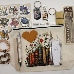 May include: An assortment of book-related items: stickers, keyrings, a bookmark, a ticket, and a canvas bag with a floral and book design. The bag contains a pen, a heart-shaped patch, and a small light.