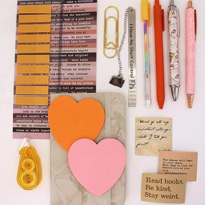 May include: A selection of stationery items, including pens, sticky notes, and a notebook. The pens are in various colours, including orange and pink, with decorative designs. There are also inspirational quote cards and a bookmark that reads "I Have No Shelf Control."
