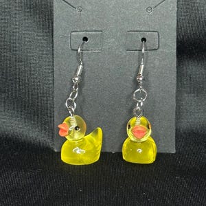 May include: A pair of yellow duck earrings with red beaks and silver-colored hooks. The earrings are made of a translucent material and are displayed on a black card.