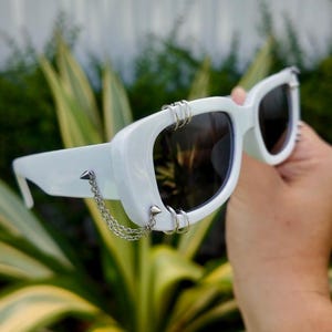 May include: White sunglasses with dark lenses, featuring silver metal accents. The sunglasses have a thick frame with a chain and spike details. The design includes multiple silver rings on the frame, adding a unique, edgy style.