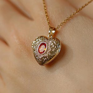 May include: A gold-colored heart-shaped pendant necklace with a small red gemstone in the center. The pendant has an ornate, detailed design. The necklace is worn around the neck.