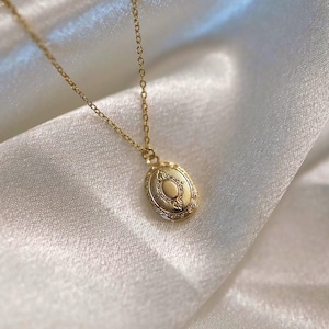 May include: A gold-colored necklace with a small, oval locket pendant. The locket has an ornate, engraved design and hangs from a delicate chain. The background is a soft, white fabric.