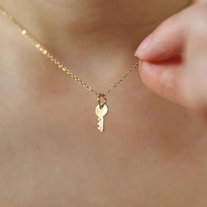May include: A delicate gold-colored necklace featuring a small key pendant. The key charm hangs from a fine chain, creating a minimalist and elegant design. The necklace is shown against a skin-toned background, highlighting the jewelry's subtle shine.