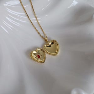 May include: A gold heart-shaped locket necklace. The locket is open, showing a small red gemstone. The necklace chain is delicate and gold. The locket is on a white surface.