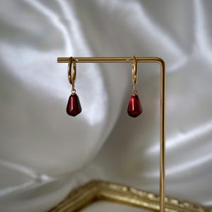 May include: Gold hoop earrings with dark red, teardrop-shaped pendants. The earrings are presented on a gold stand, set against a white fabric background. The pendants have a glossy finish.