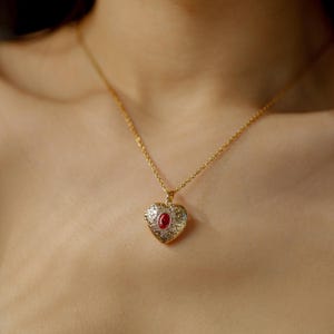 May include: A gold heart pendant necklace with a red gemstone. The heart-shaped pendant features an engraved design and hangs from a delicate gold chain. The necklace is worn on a person's chest.