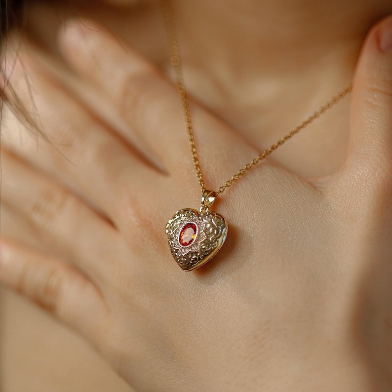 May include: A gold-colored heart-shaped pendant necklace with a small red gemstone in the center. The pendant has an ornate, detailed design. The necklace is worn around the neck.
