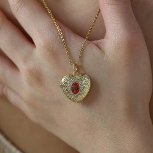 May include: Gold-coloured heart-shaped locket necklace with a red gemstone centre. The locket features detailed filigree and small clear stones. The necklace chain is delicate and gold-coloured, creating a classic jewellery piece.