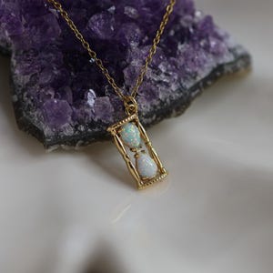 May include: A gold-toned necklace featuring a pendant shaped like an hourglass. The pendant contains two teardrop-shaped opals, one in the top chamber and one in the bottom chamber, suspended from a delicate gold-toned chain.