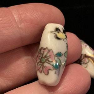 May include: White ceramic bead with a floral design in pink, green, and gold. The bead features a small bird with black and yellow accents.