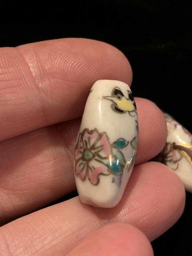 May include: White ceramic bead with a floral design in pink, green, and gold. The bead features a small bird with black and yellow accents.