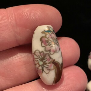 May include: White ceramic bead with a floral design in pink, gold, and green. The bead is oval shaped and has a glossy finish.