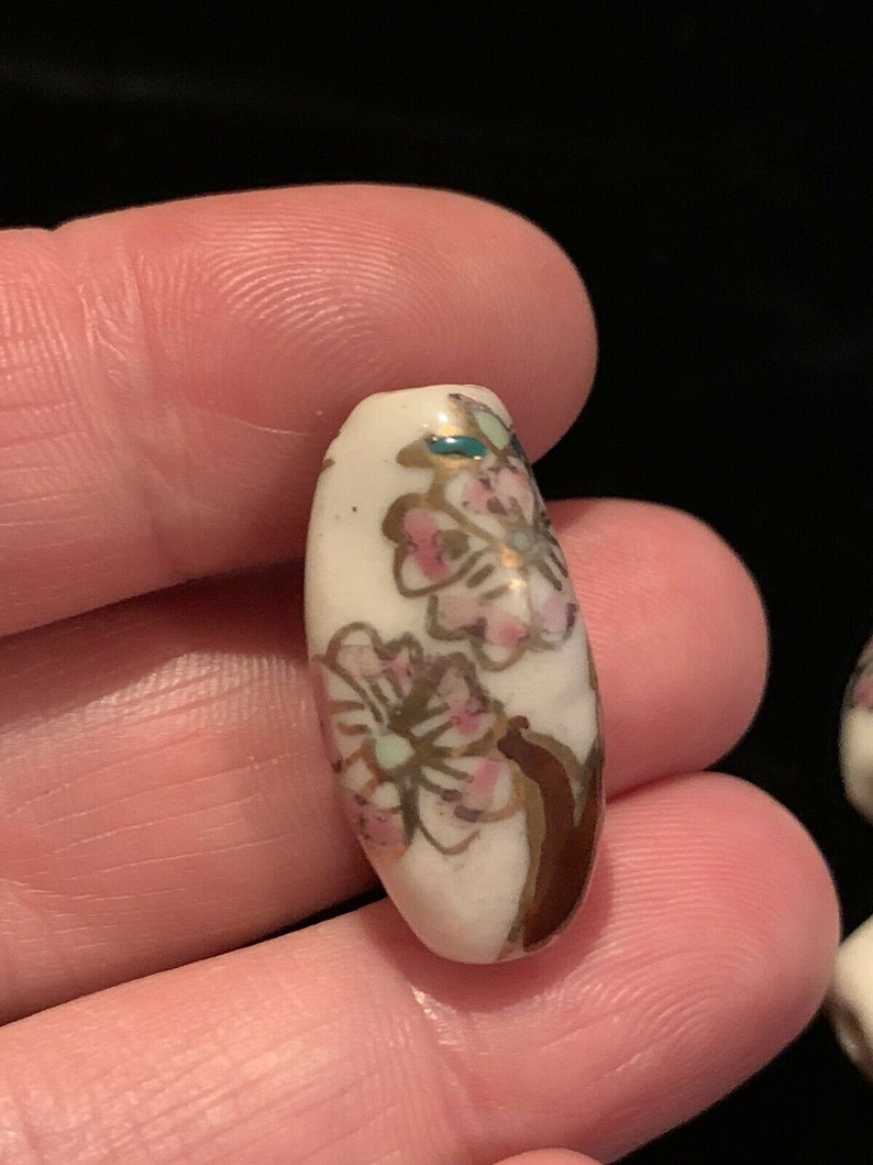 May include: White ceramic bead with a floral design in pink, gold, and green. The bead is oval shaped and has a glossy finish.