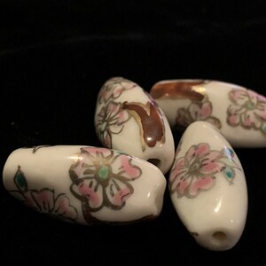 May include: Four white ceramic beads with pink and gold floral designs. The beads are oval shaped and have a glossy finish.