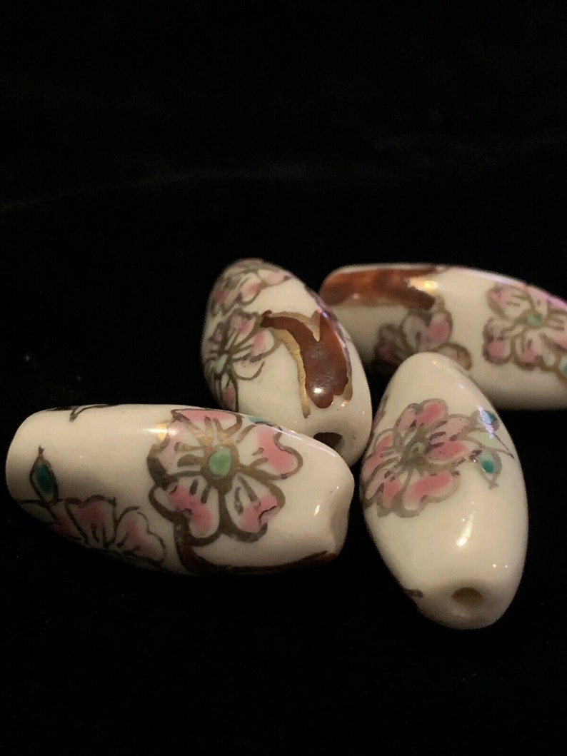 May include: Four white ceramic beads with pink and gold floral designs. The beads are oval shaped and have a glossy finish.