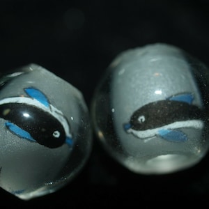 May include: Two clear glass marbles with painted black and white dolphins inside. The dolphins have blue fins and tails.