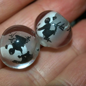 May include: Two clear glass beads with black and white silhouettes of figures inside. The figures are stylized and appear to be dancing or playing music.