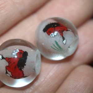 May include: Two clear glass beads with a red and black horse design painted on them. The beads are round and have a hole in the center.