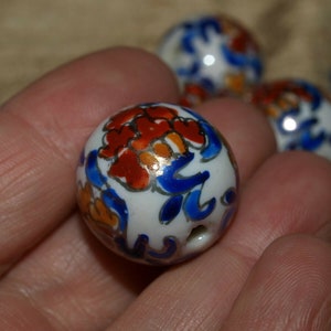 Puede incluir: Cuentas redondas de cerámica blanca con diseños florales pintados a mano en rojo, azul y naranja. Cada cuenta tiene un pequeño agujero para ensartar. La imagen muestra varias cuentas, una de ellas sostenida en una mano.