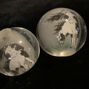 May include: Two clear glass paperweights with black and white ink drawings of horses inside. The horses are in different poses, one is rearing up and the other is standing with its head down.