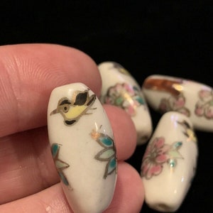 May include: Five white ceramic beads with floral and bird designs. The beads are oval shaped and have gold accents.