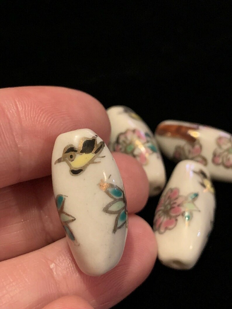 May include: Five white ceramic beads with floral and bird designs. The beads are oval shaped and have gold accents.