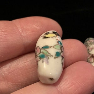 May include: White ceramic bead with a floral design in pink, green, and gold. The bead is oval shaped and has a hole in the center.