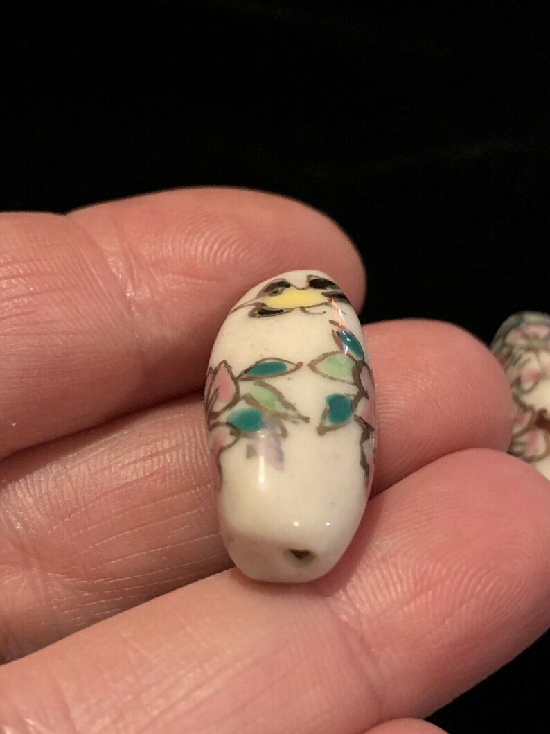 May include: White ceramic bead with a floral design in pink, green, and gold. The bead is oval shaped and has a hole in the center.