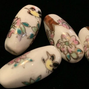 May include: Four white ceramic beads with floral and bird designs in pink, yellow, green, and gold paint.