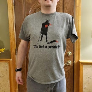 May include: A grey t-shirt with a graphic of a black knight holding a sword, with the text "'Tis but a scratch!" The knight is standing over a severed arm. The shirt is worn with grey jeans.