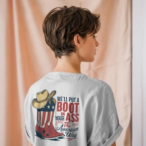 May include: A light gray t-shirt featuring a graphic of a cowboy boot, a hat, and the text "We'll put a boot in your ass the American way." The boot is red, white, and blue, with a star design.
