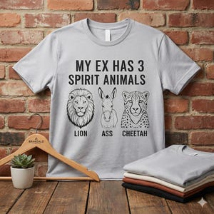 May include: Light gray t-shirt with the text "MY EX HAS 3 SPIRIT ANIMALS" above illustrations of a lion, a donkey, and a cheetah. The words "LION", "ASS", and "CHEETAH" are below the images. A wooden hanger and folded shirts are in the image.