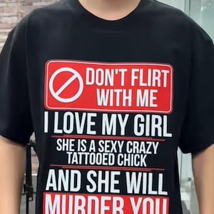 Może przedstawiać: Czarny t-shirt z odważnym wzorem graficznym. Tekst brzmi: "DON'T FLIRT WITH ME I LOVE MY GIRL SHE IS A SEXY CRAZY TATTOOED CHICK AND SHE WILL MURDER YOU" w białych i czerwonych literach.