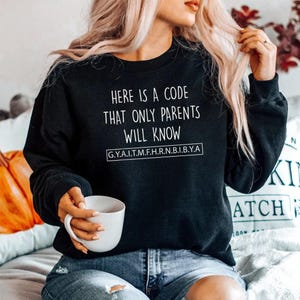May include: Black sweatshirt with white text: "HERE IS A CODE THAT ONLY PARENTS WILL KNOW" and a line of letters and numbers. A person holds a white mug. The sweatshirt is worn with blue jeans.