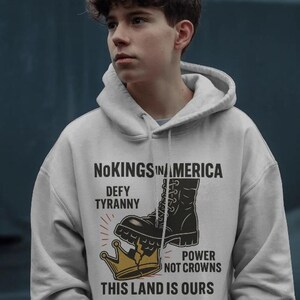 Op de afbeelding: Een lichtgrijze hoodie met een grafisch ontwerp. Het ontwerp toont een zwarte laars die op een gouden kroon trapt, met de tekst "No Kings in America, Defy Tyranny, Power Not Crowns, This Land Is Ours."