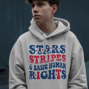 May include: A light gray hoodie with the text "STARS STRIPES & BASIC HUMAN RIGHTS" in blue and red. The design features stars and stripes, with a retro font. The hoodie has a drawstring hood.