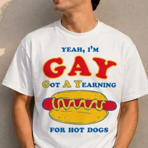 May include: White t-shirt with a graphic of a hot dog and the text "Yeah, I'm Gay, Got a Yearning for Hot Dogs" in red, yellow, and blue. The shirt is a casual, unisex style.
