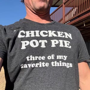Può includere: T-shirt grigio scuro con testo bianco che recita "CHICKEN POT PIE three of my favorite things". La maglietta a maniche corte è realizzata in un materiale morbido. La persona che indossa la maglietta è all'aperto.