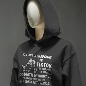 Può includere: Felpa con cappuccio nera con testo bianco e grafica di unicorno. Il testo recita: "No, non posso usare Snapchat o TikTok, ma posso scrivere in corsivo, fare matematica senza calcolatrice e dire l'ora su un orologio con le lancette."
