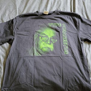 May include: Dark gray t-shirt featuring a green-tinted image of a man wearing glasses and a mustache. The text "TYPE 2 DIABETUS" is printed in green on the shirt. The shirt is made of a soft material.