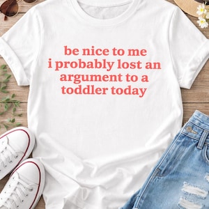 May include: White t-shirt with the text "be nice to me i probably lost an argument to a toddler today" in red. The shirt is paired with denim shorts, white sneakers, sunglasses, and a straw hat.