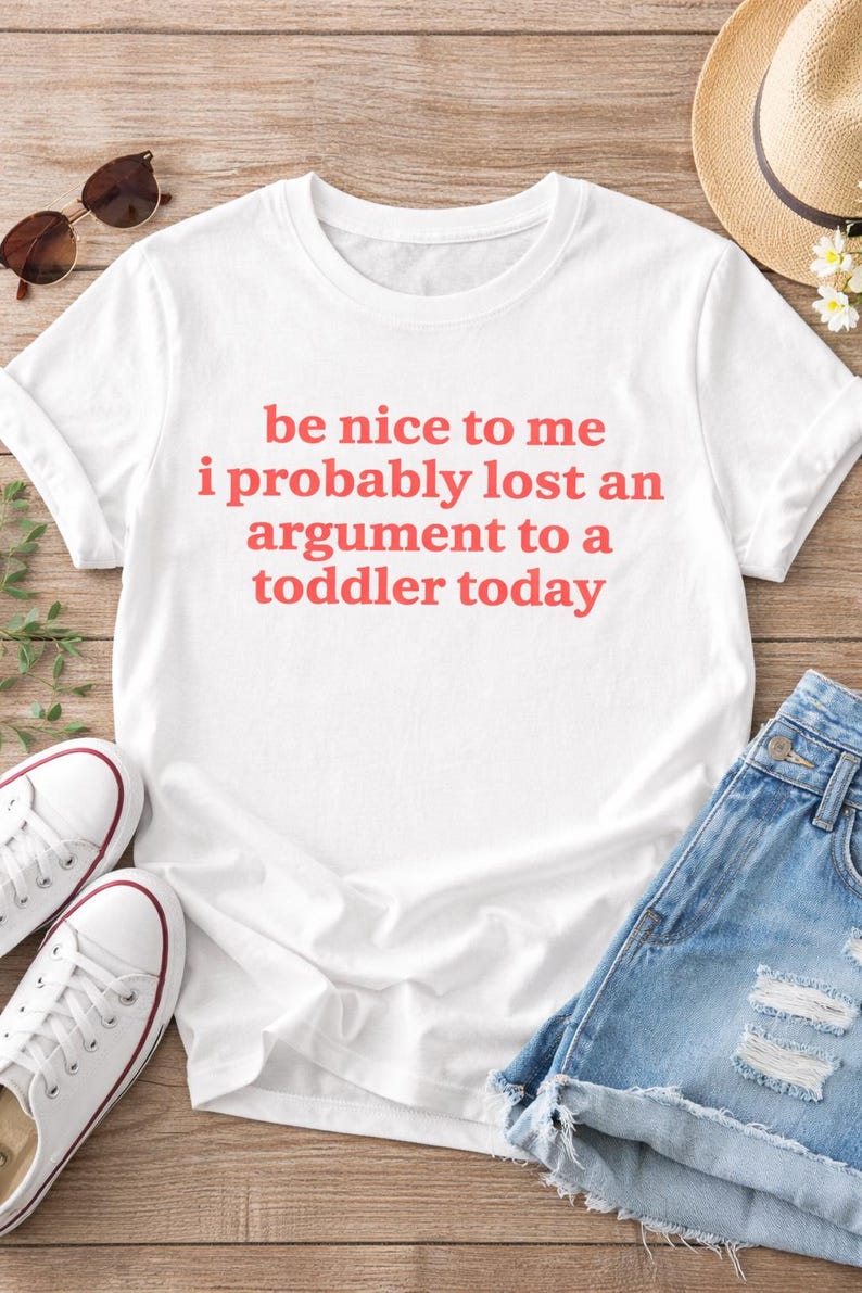 May include: White t-shirt with the text "be nice to me i probably lost an argument to a toddler today" in red. The shirt is paired with denim shorts, white sneakers, sunglasses, and a straw hat.