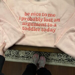 May include: A light pink sweatshirt with the embroidered text "be nice to me i probably lost an argument to a toddler today" in a coral color. The sweatshirt is on a dark wooden surface.