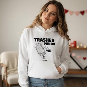 May include: White hooded sweatshirt with a front pocket. It features a black graphic of a raccoon and the words "TRASHED PANDA". The hoodie also has the word "#RaccoonLife" printed below the graphic. The model is wearing blue jeans.