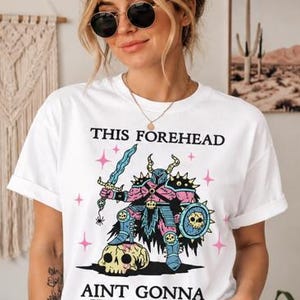 May include: A white t-shirt featuring a graphic illustration of a pink and blue armored warrior standing on a skull, holding a sword and shield. The text reads THIS FOREHEAD AINT GONNA KISS ITSELF, surrounded by small pink star icons on the front.