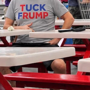 May include: A gray t-shirt with the text "TUCK FRUMP" in red and blue block letters. The person wearing the shirt is seated at a white table with red legs. A black knife rests on the table.