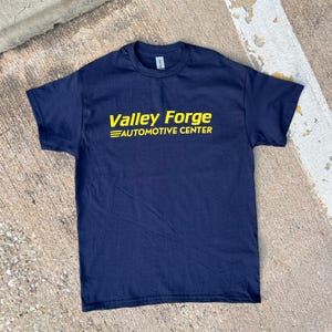 May include: Navy blue t-shirt with the text "Valley Forge AUTOMOTIVE CENTER" in yellow. The shirt is a crew neck style and appears to be made of cotton. The shirt is laying on a concrete surface.