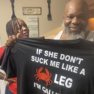 Puede incluir: Camiseta negra con el texto "IF SHE DON'T SUCK ME LIKE A LEG I'M CALLIN MY" y un gráfico de cangrejo rojo. La camiseta también presenta un logotipo de "OLD BAY". La imagen la sostienen dos personas.