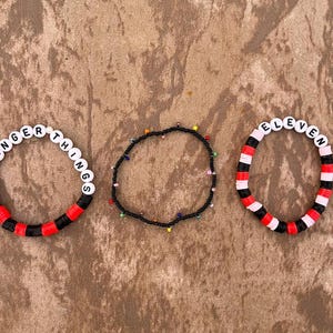 May include: Three beaded bracelets. One bracelet spells "STRANGER THINGS" in white beads with black and red accents. Another is a black beaded bracelet with colorful beads. The third bracelet spells "ELEVEN" with red, black, and white accents.